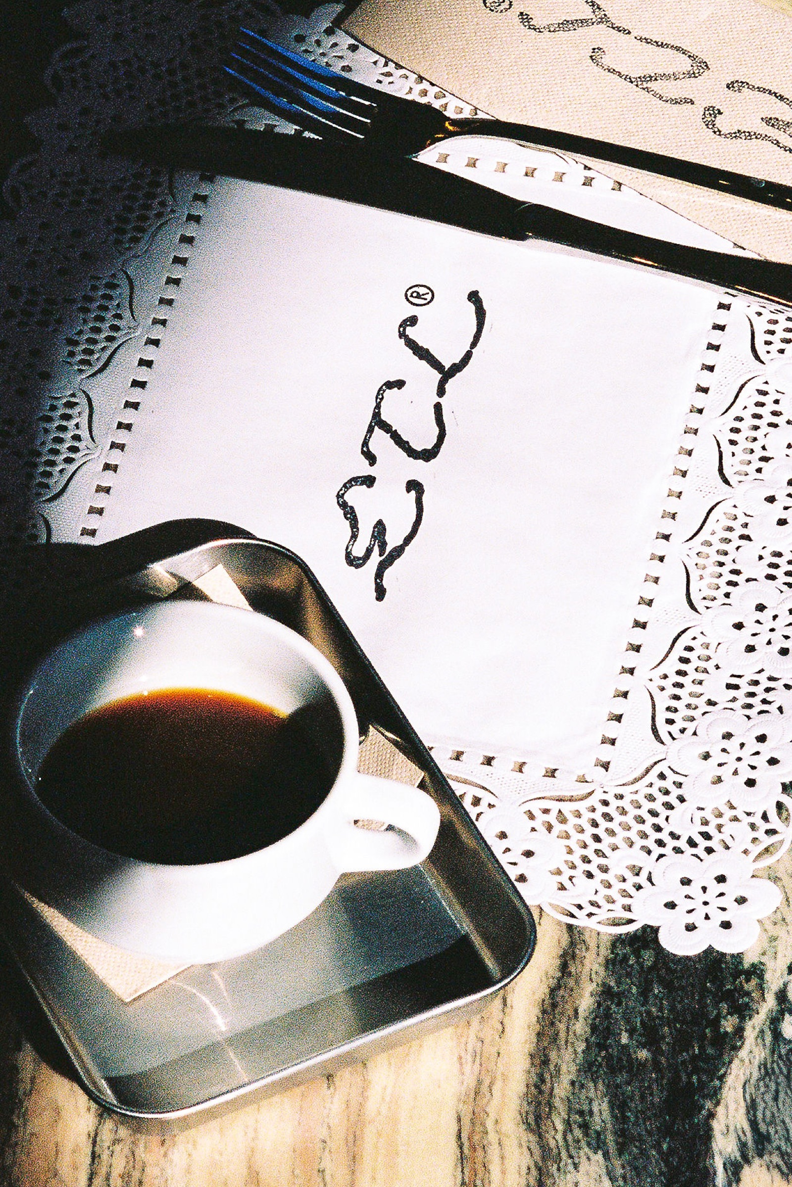 Taza de café espresso sobre bandeja de acero junto a servilleta blanca con la marca cursiva ETL bordada y doily de papel calado.