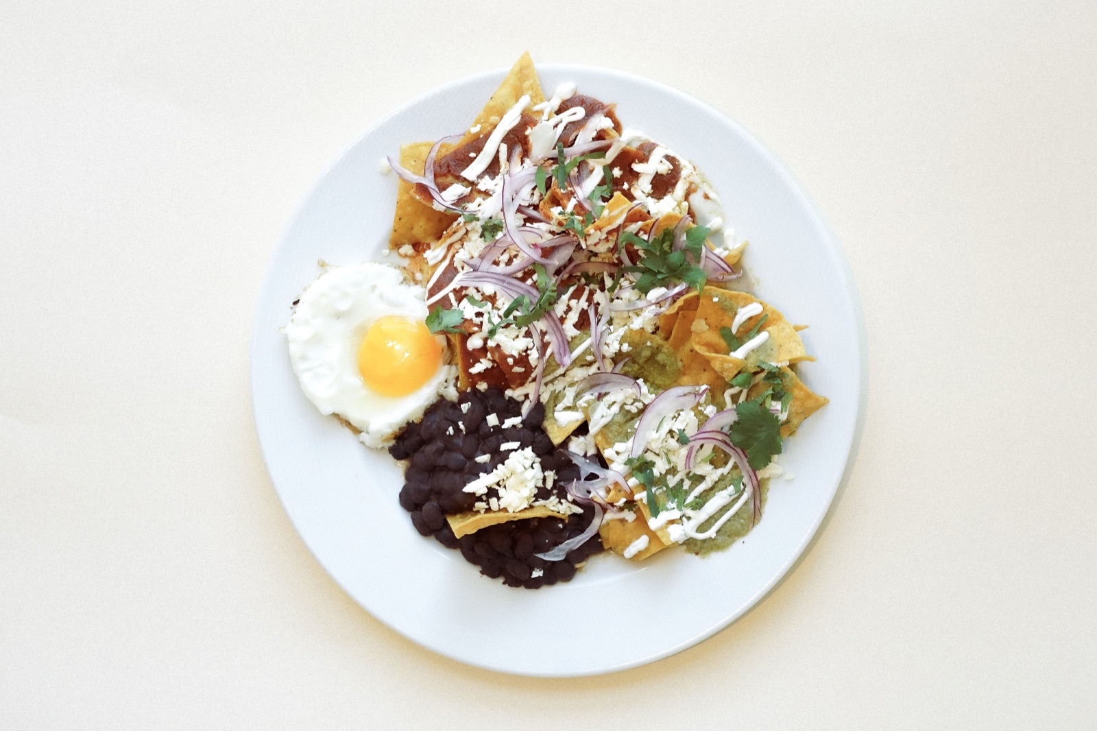 Chilaquiles con huevo frito, frijoles negros, cebolla morada, cilantro y queso desmoronado, vista cenital.