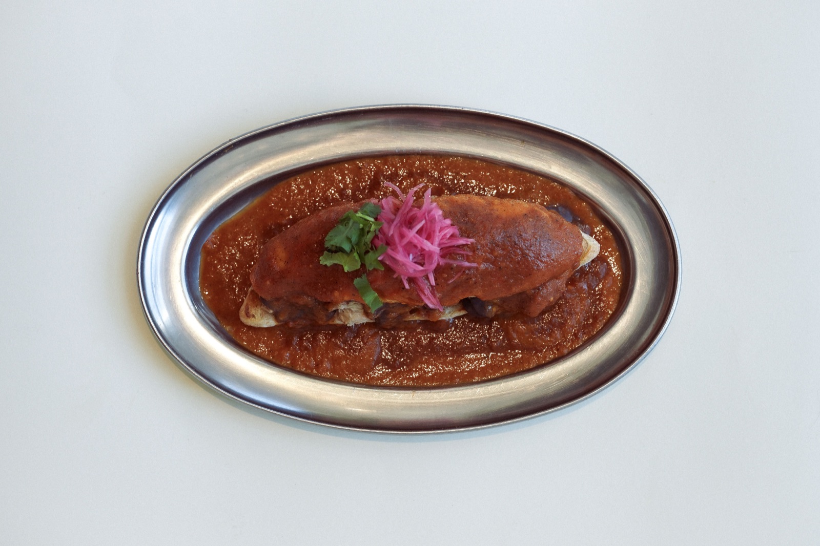 Mollete bañado en salsa roja profunda con cebolla encurtida sobre bandeja oval de acero.