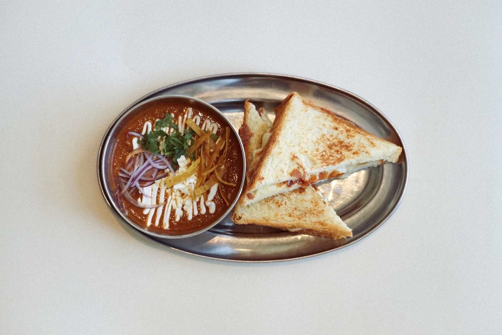 Sopa de tortilla en cazuelita de acero junto a sandwich bikini cortado por la mitad sobre bandeja oval.