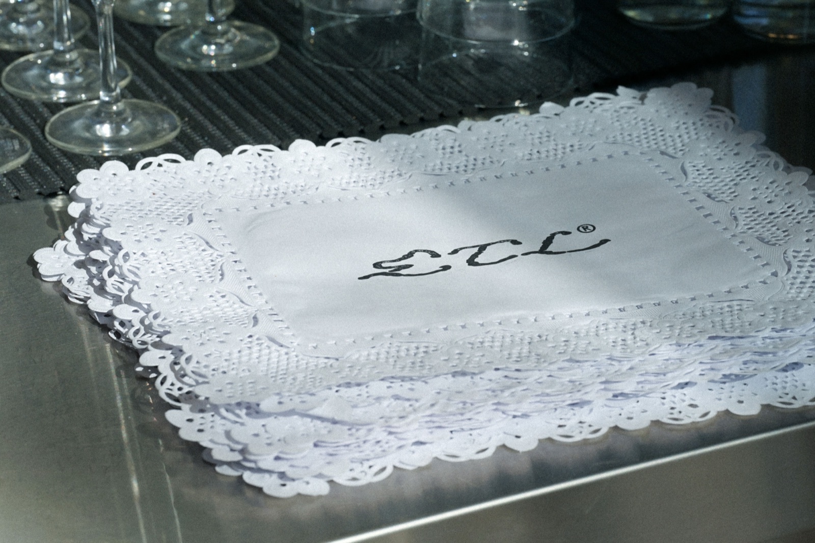 Servilleta blanca con la marca cursiva ETL® bordada sobre doily de papel calado, copas al fondo.